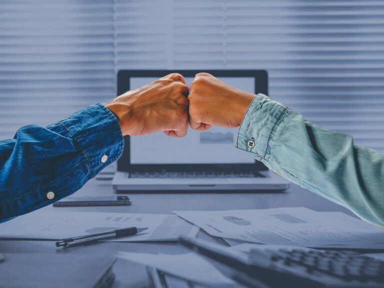 Two hands fist-bumping in front in front of desk with computer